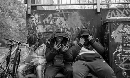 Three hooded individuals sit, faces concealed, by "THE ST" graffiti.