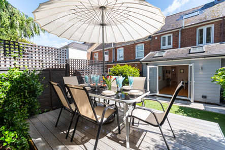 Outdoor dining area with table, chairs, umbrella, decking, house background, sunny.