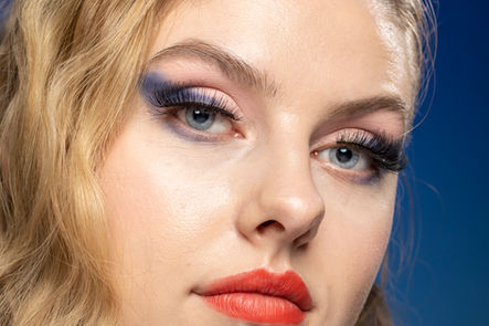 Woman's face with dramatic blue eyeshadow, long lashes, orange-red lipstick.