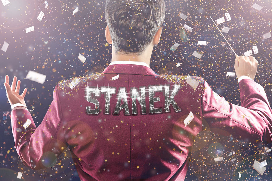 a conductor with his back to us raises his baton in the air as confetti pours down all around; across his shoulders: STANEK