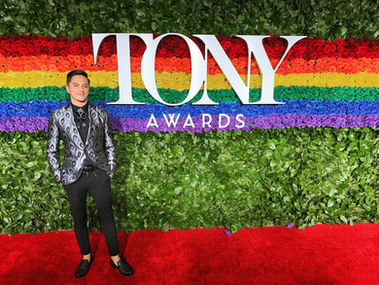 Joseph Stanek at the Tony Awards red carpet, dressed in a silver patterned blazer, standing before a rainbow floral backdrop.