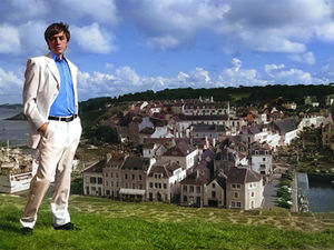 Joseph Stanek stands on the ancient walls of Le Citadelle, overlooking a picturesque French coastal paradise; he wears a white suit and a blue button-down; cast member of Lyrique en Mer's La Traviata production