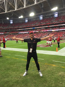 Joseph Stanek, wearing silver shoes, black jeans, a black Arizona Cardinals' polo, and a backwards Cardinals' hat, stands on the field with arms outstretched