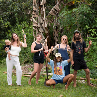 A small group of adults and a child standing together on grass in a lush outdoor setting, holding sticks and tools, celebrating community, teamwork, and connection to the land at Hamakua Sanctuary. If you want this to lean more playful, more reverent, or more focused on the residency/volunteer aspect, I can tailor it exactly to that tone.