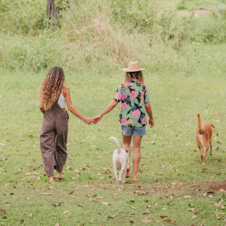 Two people holding hands while walking through a grassy field with two dogs, surrounded by open green landscape, reflecting connection, calm, and togetherness at Hamakua Sanctuary. If you’d like a slightly more poetic version or one that emphasizes family, stewardship, or daily life at the sanctuary, I can refine it.