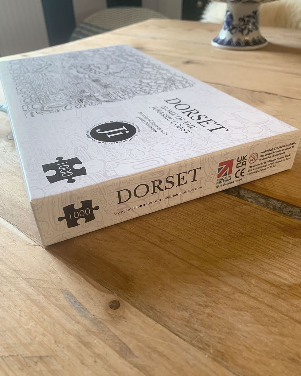 Thumbnail: Corner of a Dorset jigsaw puzzle box by James Illustrates on a wooden dining table.