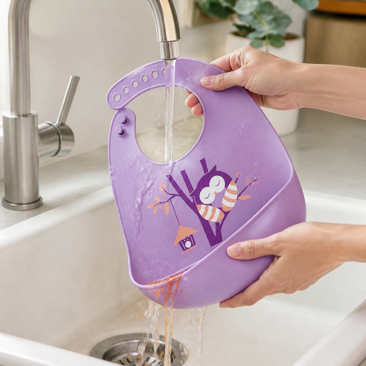 Adult rinsing the Purple Owl silicone bib and its food-catching pocket in a sink