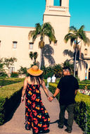 Couple holding hands walking toward a large building with palm trees.