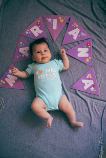 Baby with MARIANA banner, wearing onesie on blanket.