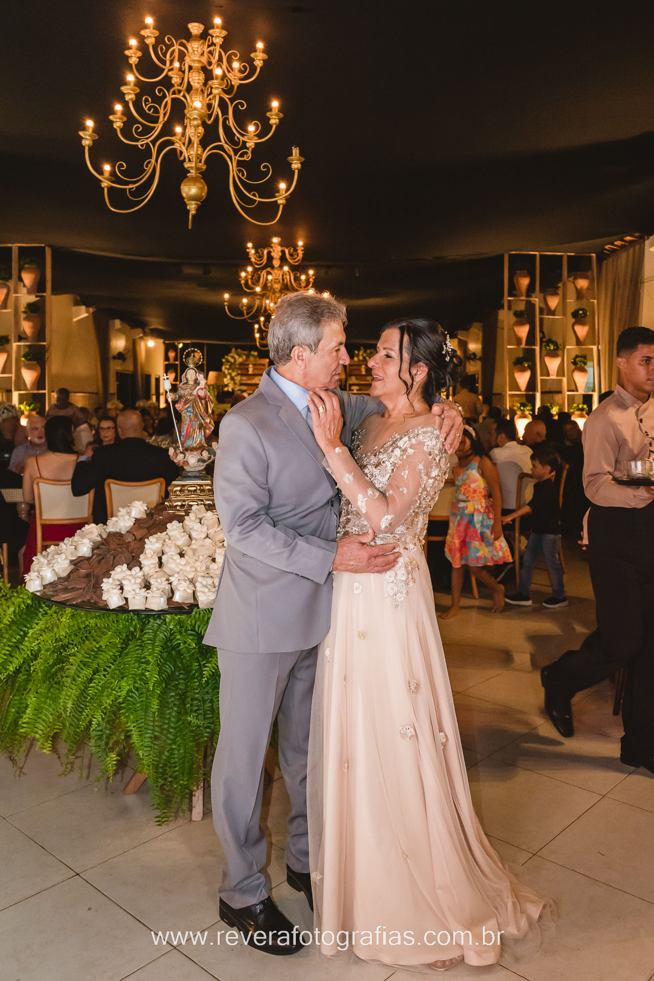 Foto do casal junto em festa de bodas de ouro. Ao lado da mesa de doces