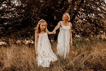 Mother and daughter hold hands in white dresses and walk through tall grass