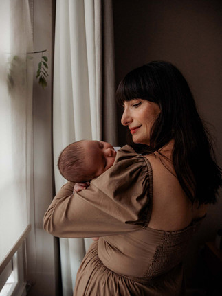 Mum holds newborn baby in brown dress next to window