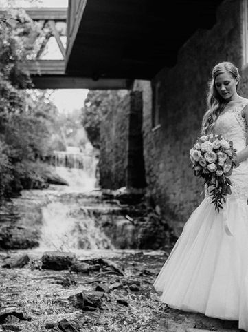 Elegant black and white bridal portrait of a bride standing by a historic stone wall with a rushing natural waterfall and overhead bridge in the background at Ancaster Mill.