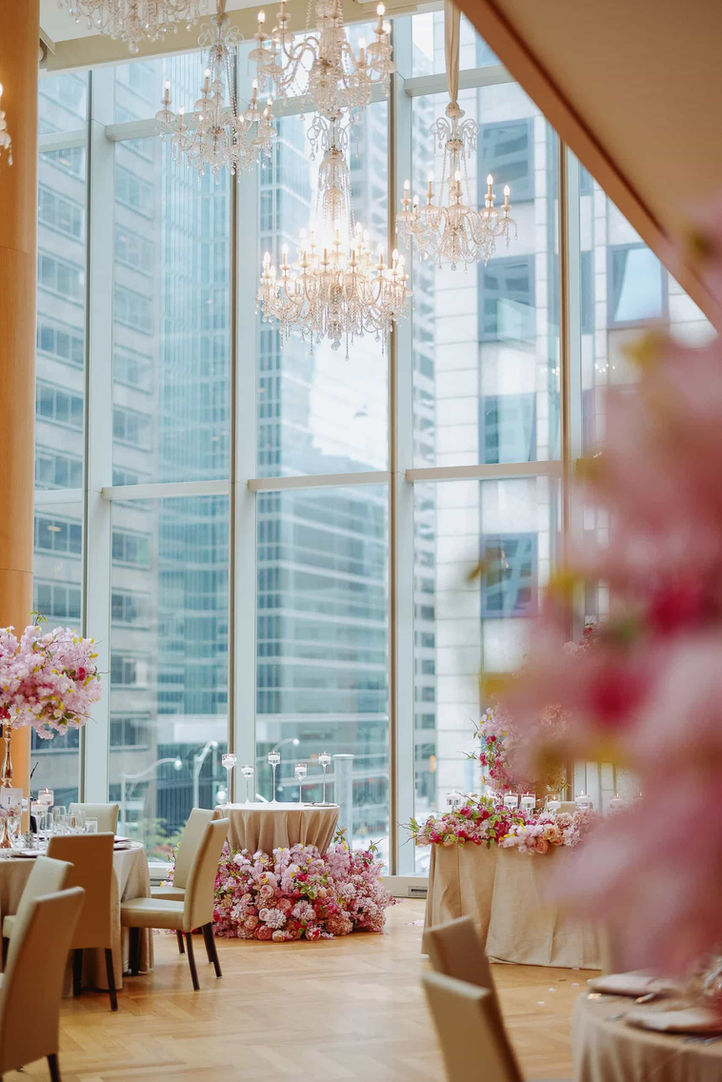 A view inside a wedding reception venue with large windows overlooking a city skyline. Two round tables are set with tablecloths and chairs, one heavily decorated with pink flowers around the base. Floral arrangements are also on the tabletops. Crystal chandeliers hang from the ceiling.