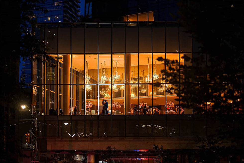 A dramatic nighttime photo of a wedding reception, with the couple silhouetted inside a modern glass venue filled with the warm glow of chandeliers.