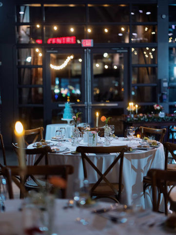 An intimate, moody reception table setup featuring cross-back wooden chairs, glowing candles, and menus inside a glass-walled venue.
