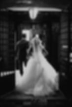 Bride in gown and man in suit run through open doors, captured in black-and-white. Light streams in, creating a dramatic, joyous scene.