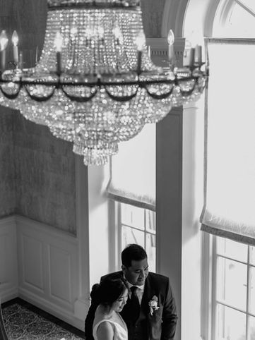 A timeless black and white photograph of a groom leaning against a window frame beneath a massive, intricate crystal chandelier.