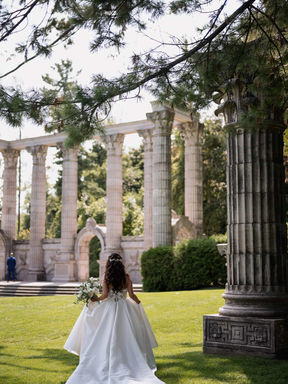A magnificent back view of a bride with a long train walking across the lawn toward the soaring historic stone pillars at Guild Park.