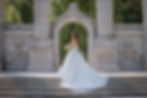 Bride in a flowing white dress holding flowers stands on stone steps, framed by ornate columns and arch, with lush greenery behind.