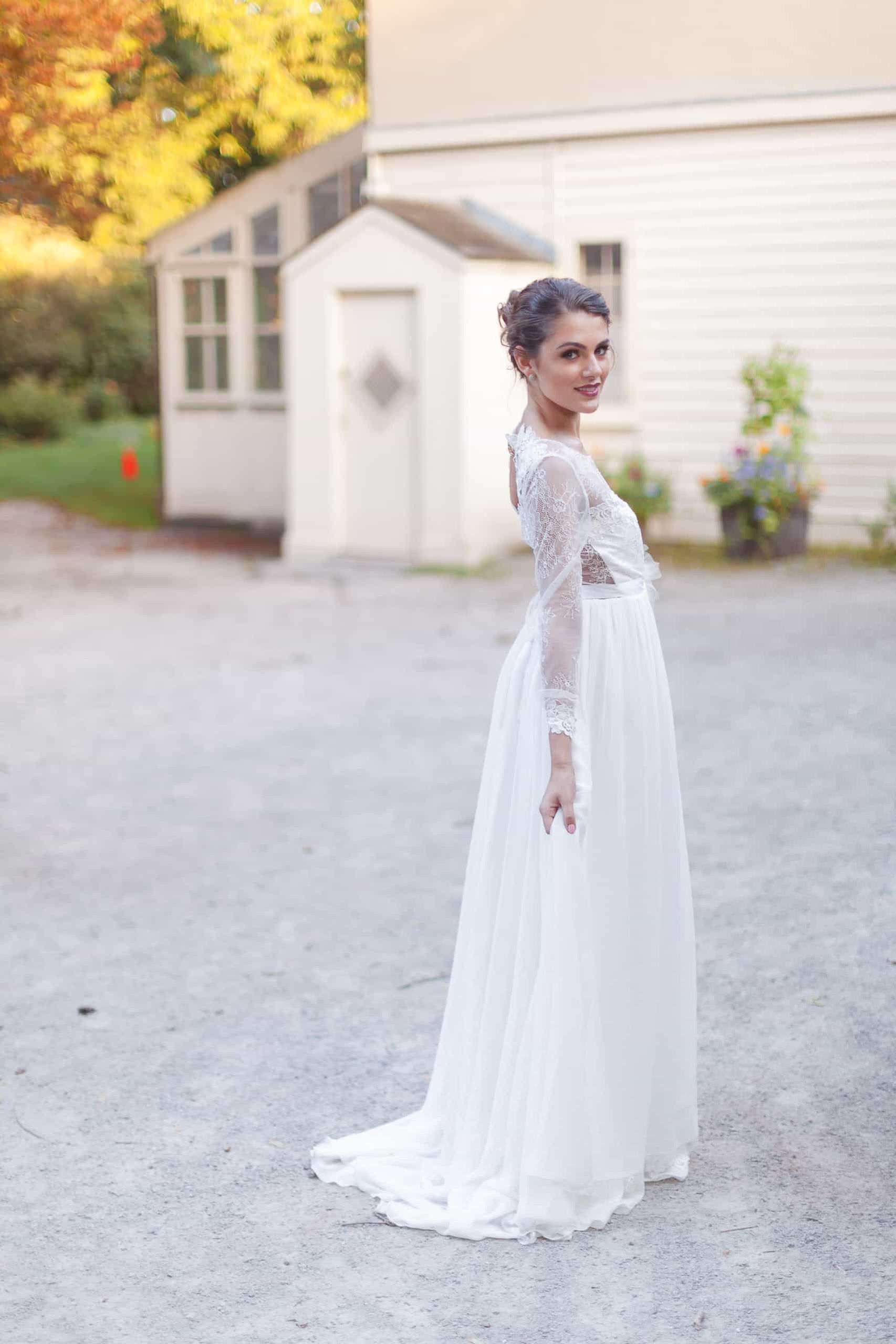 A bride in a long-sleeved A-line wedding dress posing outdoors in front of a white carriage house wedding venue.