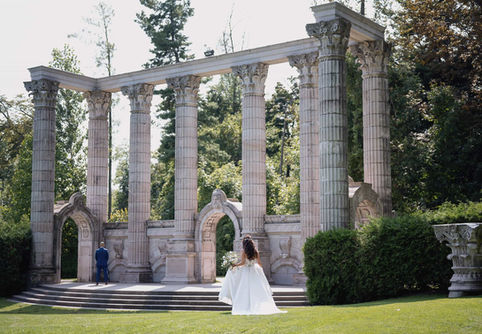 A groom in a blue suit kissing his bride while standing under a historic, vine-covered stone archway.
