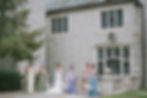 Six women in colorful dresses hold parasols, posing by a stone building. The serene scene features greenery and elegant architecture.