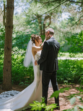 A bride and groom sharing a quiet, intimate moment on a sun-dappled forest pathway surrounded by lush woodland greenery.
