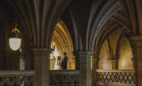 A creative, symmetrical architectural portrait of a bride and groom standing on a balcony inside a stunning gothic stone corridor with vaulted ceilings.