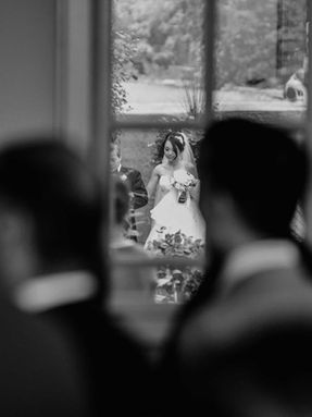 Artistic black and white photo focusing on the bride in the distance, framed by the blurred silhouettes of wedding guests.