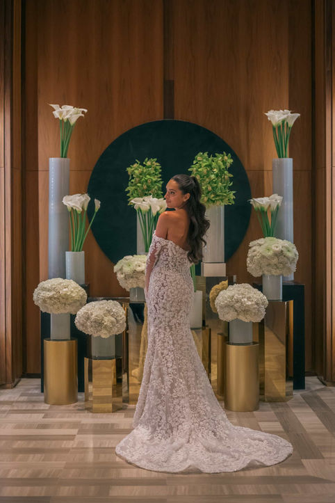 Back view of a bride displaying the train of her lace wedding dress, surrounded by modern white calla lily floral arrangements and gold pedestals.