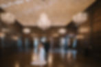 A couple walks in an elegant hall with chandeliers and wood paneling, she wears a white gown. The mood is serene and romantic.