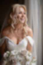 Bride in lace gown holding bouquet, looking out a window. Elegant room with a vintage chair, blue curtains, bright and serene ambiance.