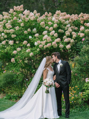 A romantic couple portrait of a bride and groom kissing in front of a massive, blooming wall of white hydrangea bushes.