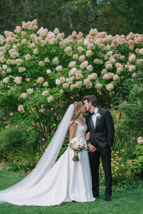 Romantic kiss shared by a bride and groom in front of a large, blooming white hydrangea bush in the estate gardens.