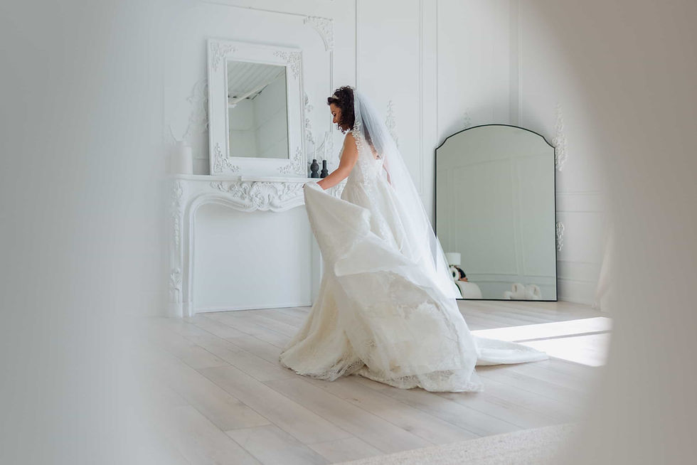 Bride in white gown and veil holds dress in bright room with ornate mirror and wooden floor, creating an elegant and serene atmosphere.