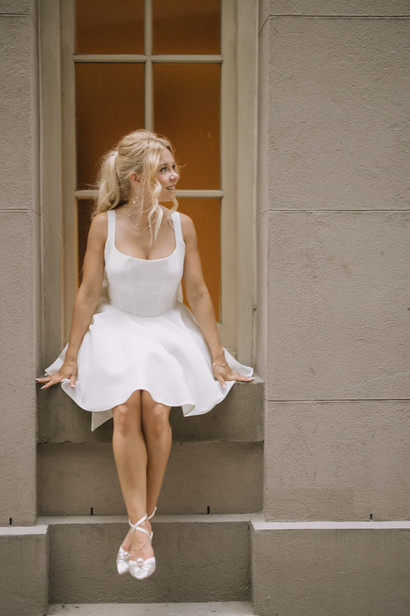 A smiling woman in a short white dress and stylish heels sits elegantly in the windowsill of a stone building during her engagement photoshoot.