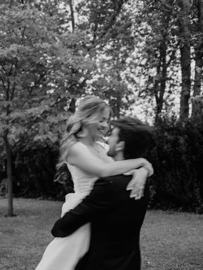 Expressive black and white candid wedding photography of a groom lifting and hugging his bride during a romantic outdoor session surrounded by tall trees.