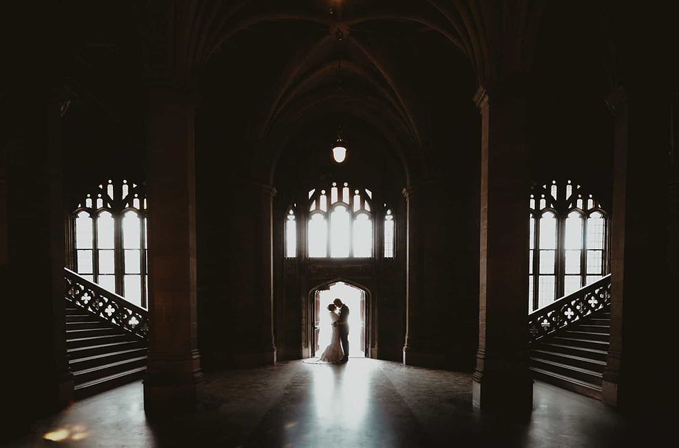 Silhouetted bride and groom embrace in a dim Gothic hall with tall windows and bright doorway light