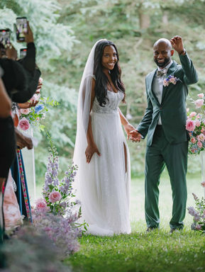 A joyful bride and groom in a green suit walking hand-in-hand down a forest aisle as guests celebrate their ceremony recessional.