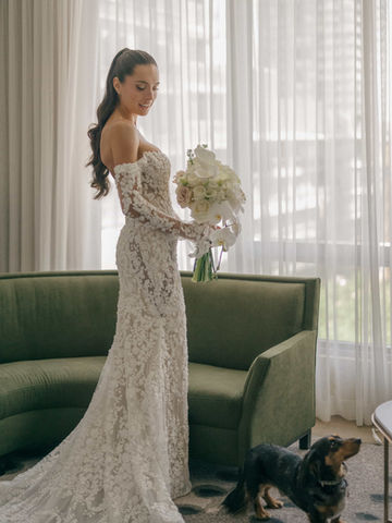 A full-length portrait of a bride wearing an off-the-shoulder lace gown, holding a white bouquet near a green velvet sofa during a Toronto wedding morning.
