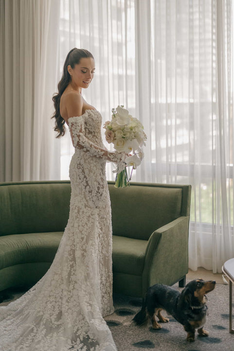 A full-length portrait of a bride wearing an off-the-shoulder lace gown, holding a white bouquet near a green velvet sofa during a Toronto wedding morning.
