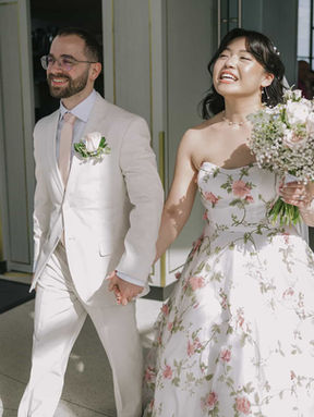 A happy couple, featuring a bride in a floral embroidered dress and groom in a white suit, laughing during their grand entrance.