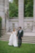 Bride and groom in elegant attire walk hand in hand on green grass, smiling against a backdrop of stone columns and lush greenery.