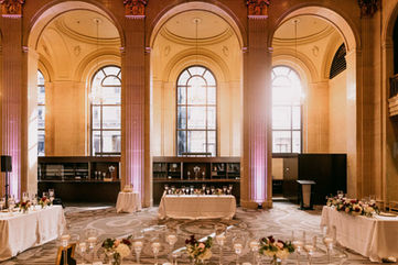 A beautiful wide shot of the Grand Banking Hall arranged for an indoor ceremony, bathed in warm natural light from massive arched windows.