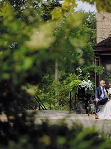 A romantic long-distance couple portrait of a bride and groom sitting on a stone bench, creatively framed through soft, out-of-focus green garden leaves.