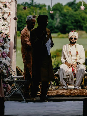 A South Asian groom seated at an outdoor mandap during a dusk wedding ceremony, surrounded by family and an officiant.
