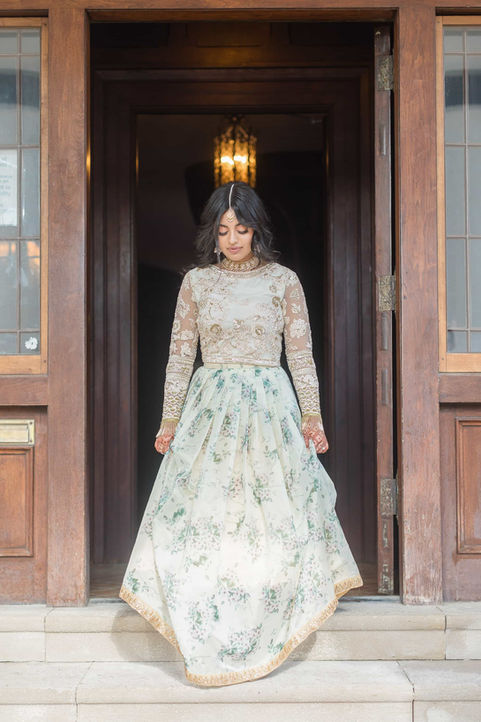 A South Asian bride standing beautifully in an ornate wood doorway wearing a pastel floral embroidered lehenga.
