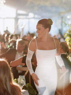 A smiling bride in a modern, long-sleeved gown walks into her bright wedding reception room as guests applaud.