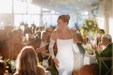 A smiling bride in a modern, long-sleeved gown walks into her bright wedding reception room as guests applaud.
